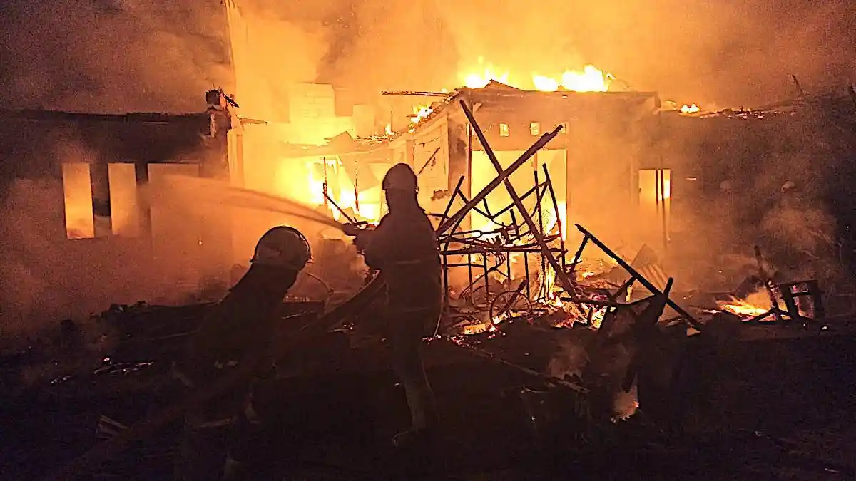 Firefighters deploying hoses to extinguish flames engulfing semi-permanent houses.